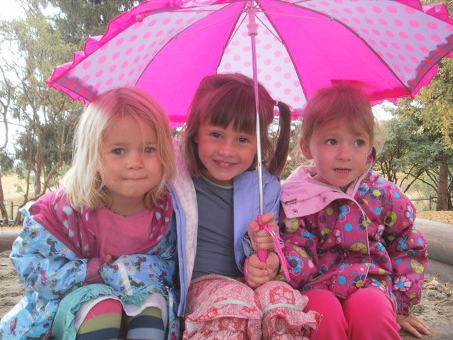 Three children sit under a pink polka-dot umbrella outdoors, wearing colorful jackets.