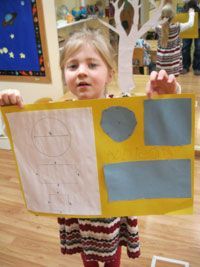 Child holding a yellow poster with blue paper shapes in a classroom setting