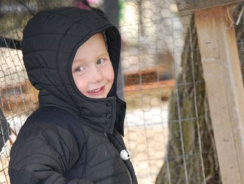 Child in a black hooded jacket smiling beside a wire fence outdoors.