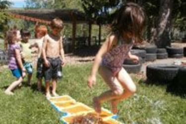 Children playing an outdoor stepping path game on grass near tires and a shaded shelter.