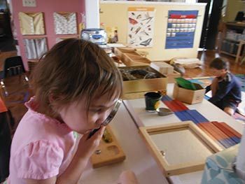 Child using a wooden toy at a classroom activity table, with another child in the background.