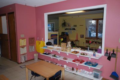 Pink room with a large serving window, shelves, and a small table in a classroom or play area