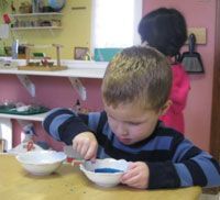 Child in a striped shirt eating from a bowl at a table in a classroom, with another child in the background.