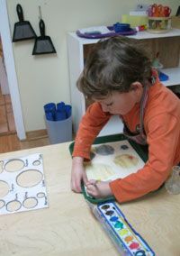 Child in an orange shirt using a paint roller on paper at a classroom table