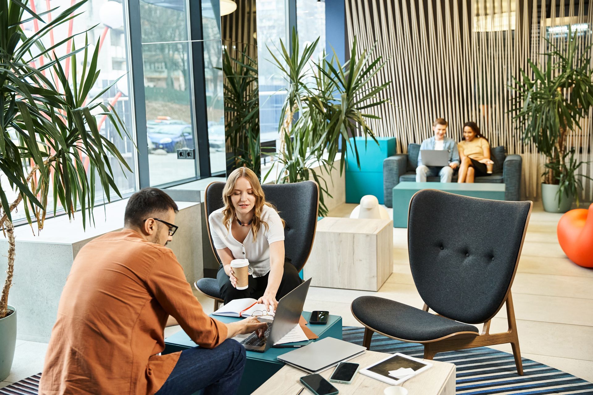 People collaborating in modern office lounge area, with laptops, plants, and colorful seating.