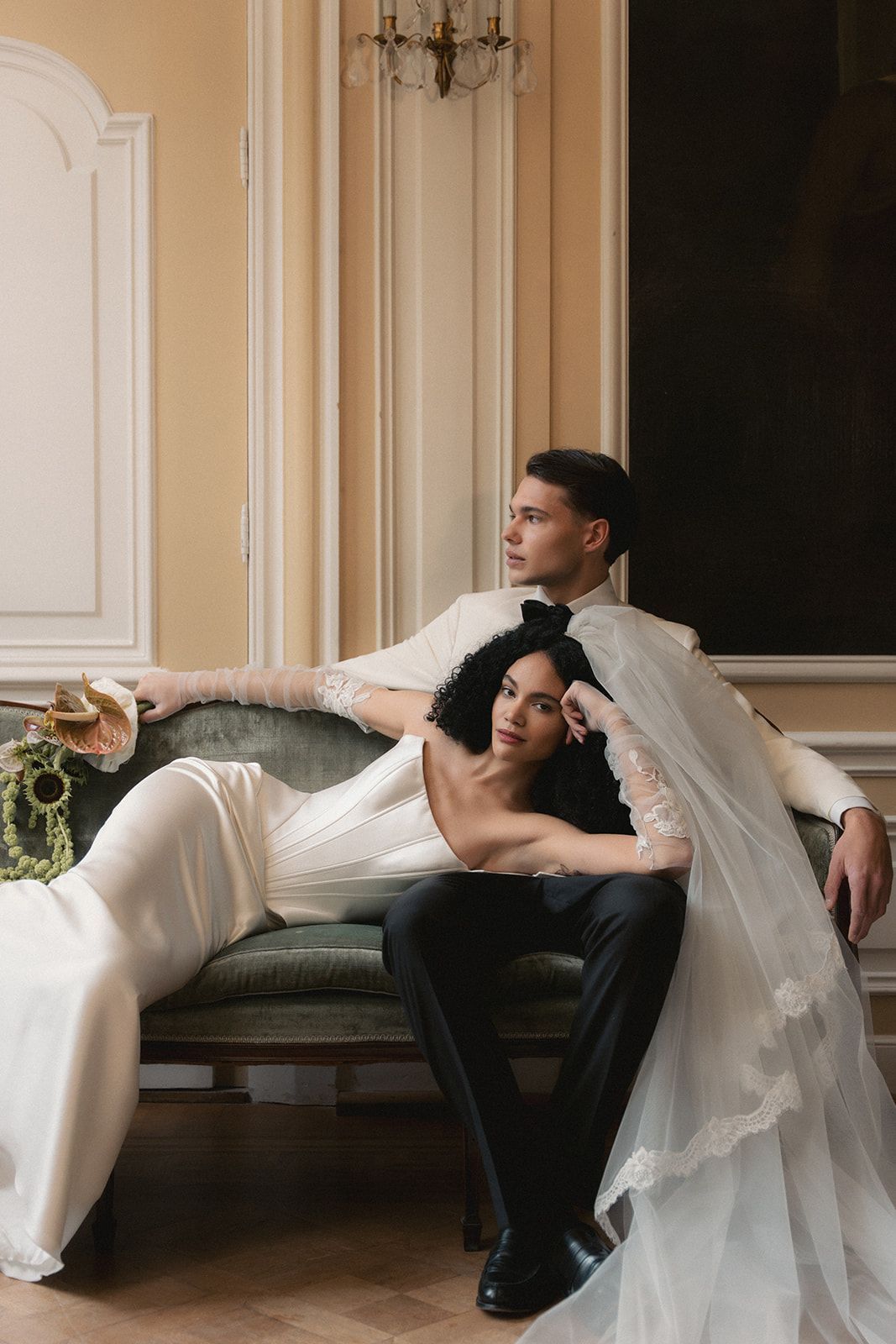 Couple in white and black seated on a sofa in an elegant room, leaning together with a sheer veil draped over them
