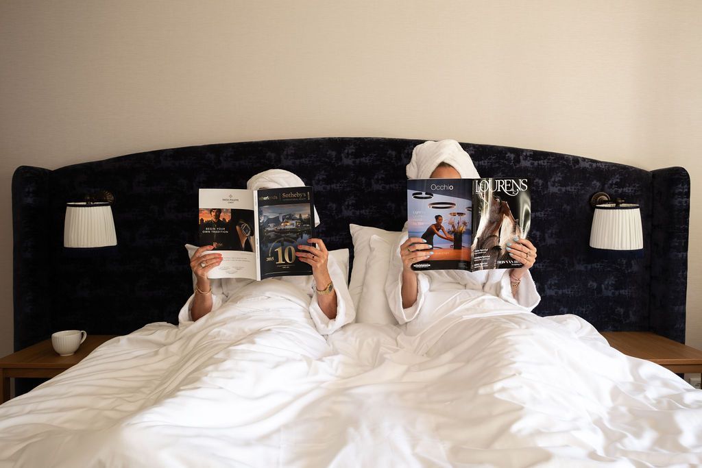 Twee mensen liggen onder witte dekens in bed en houden tijdschriften vast tegen een donker hoofdeinde met wandlampen.