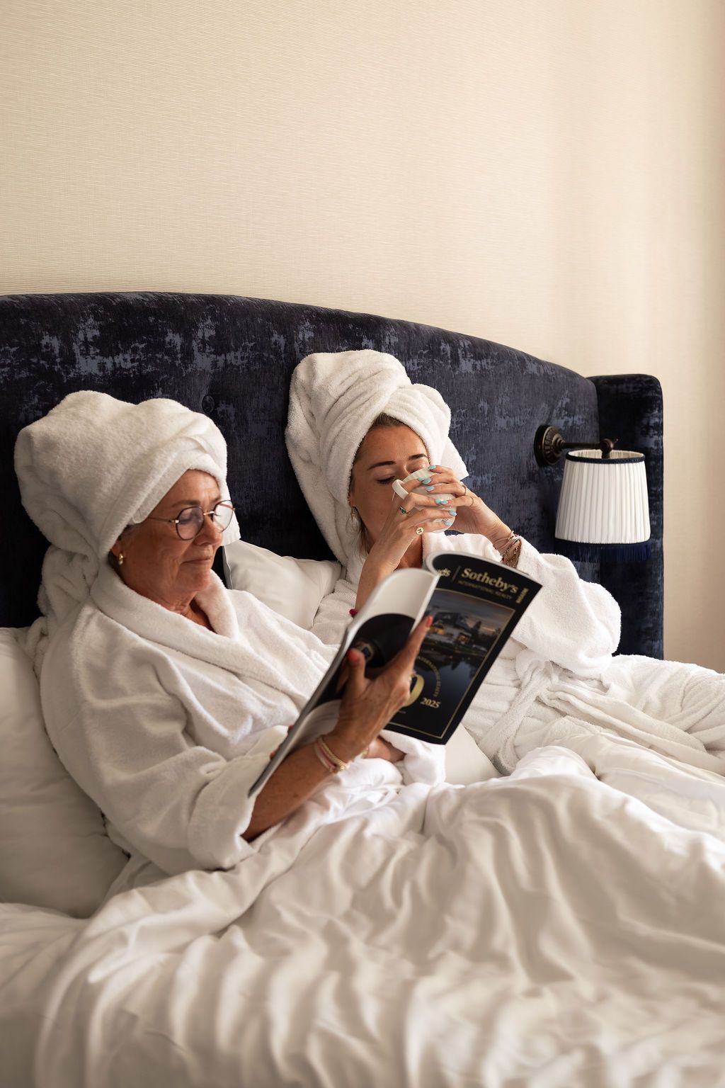 Twee mensen in witte badjassen en handdoeken zitten in bed een tijdschrift te lezen naast een lamp.