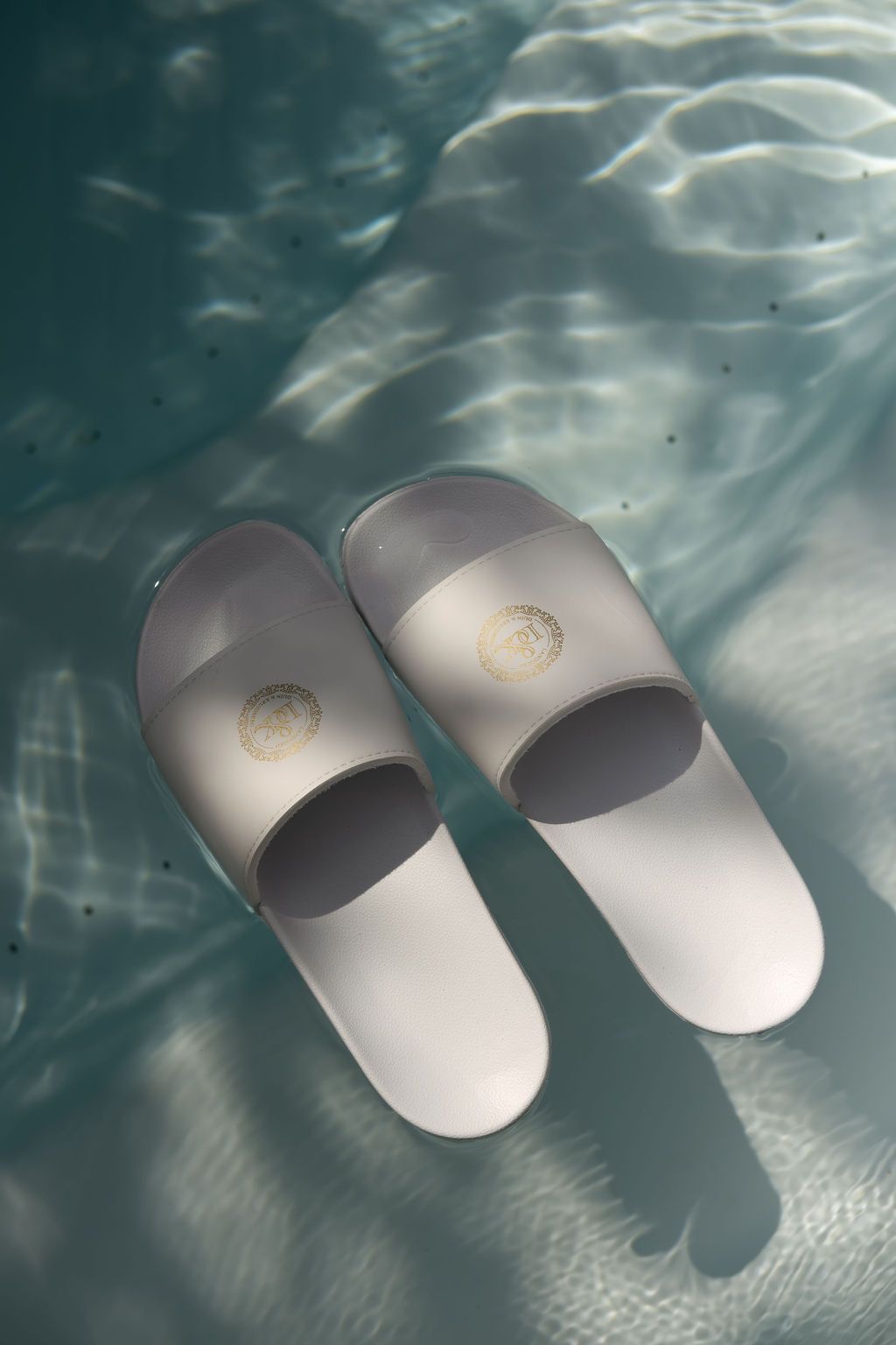 Een paar witte slippers op een zonovergoten zwembadoppervlak met rimpelende waterschaduwen.