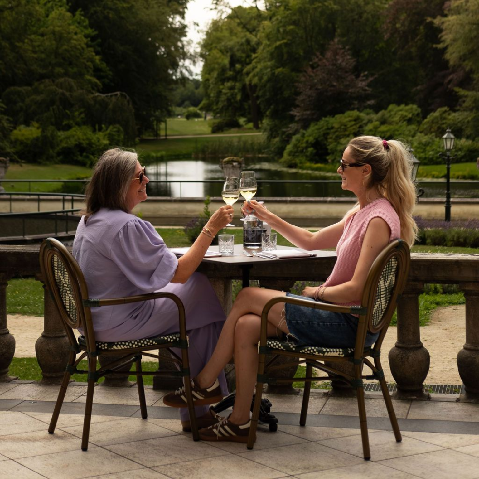 Twee vrouwen heffen het glas met wijn op een terrasje aan een vijver, met bomen en groen op de achtergrond.