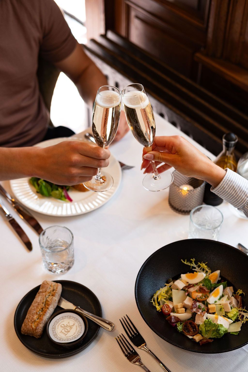 Twee mensen klinken met champagneglazen aan een restauranttafel met salade, brood en drankjes.