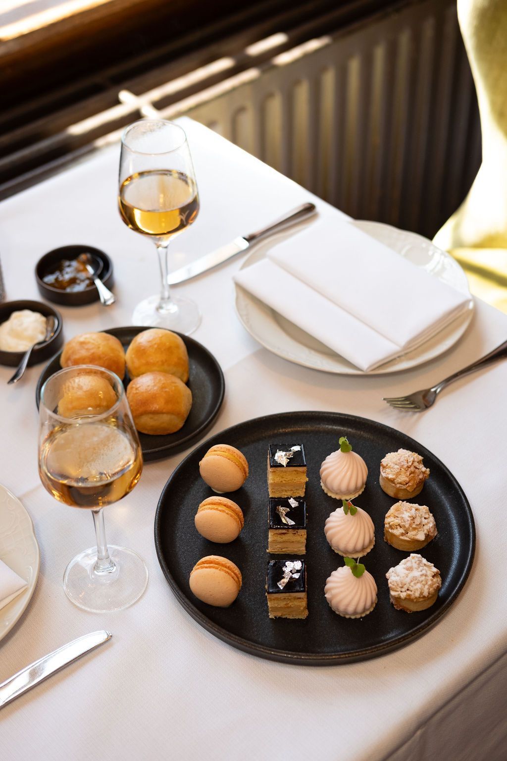 Een elegant gedekte tafel met kleine gebakjes op zwarte bordjes, wijnglazen en gevouwen witte servetten.