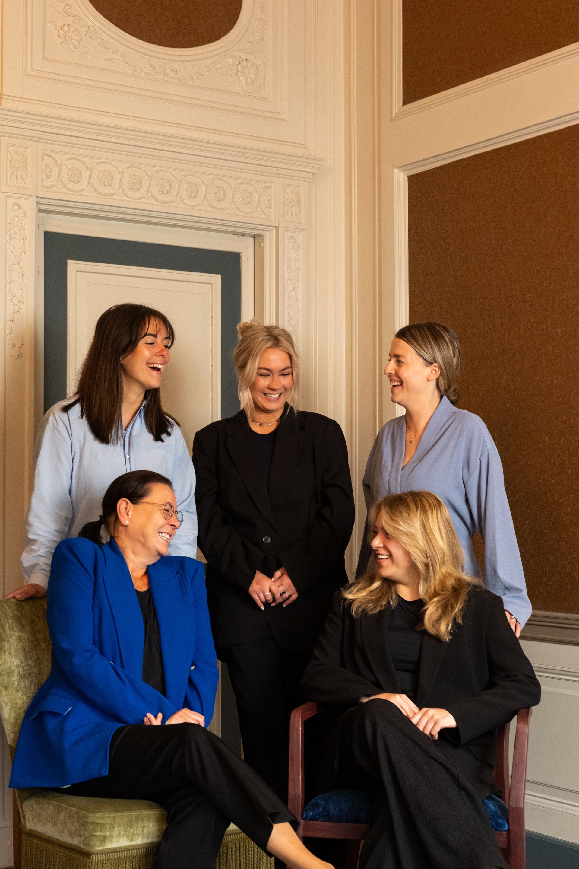 Five people posing together in a bright room with ornate walls, smiling and chatting.