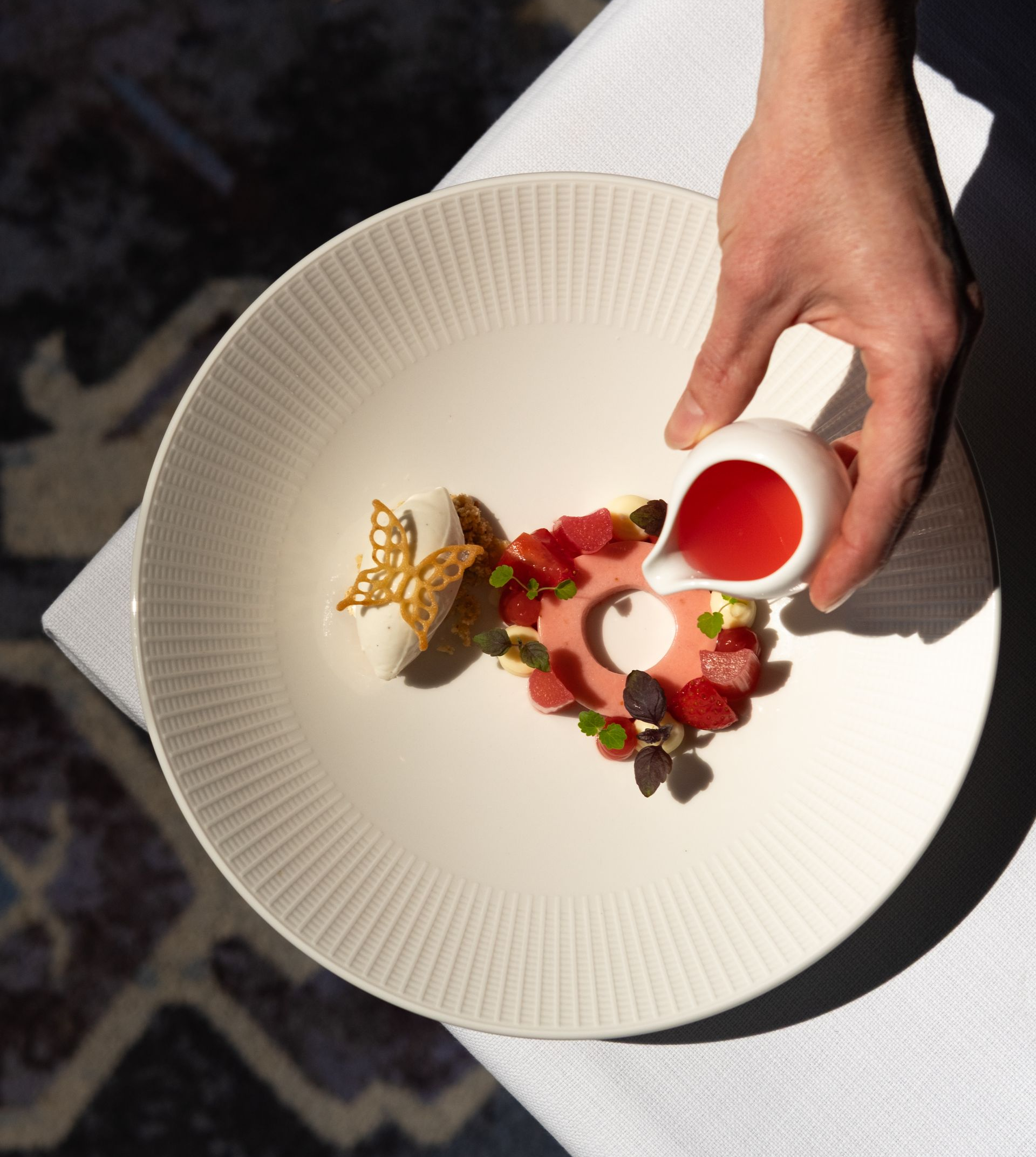 Eine Hand platziert rote Soße neben einem Dessertteller mit Erdbeeren auf einem weißen, strukturierten Teller.