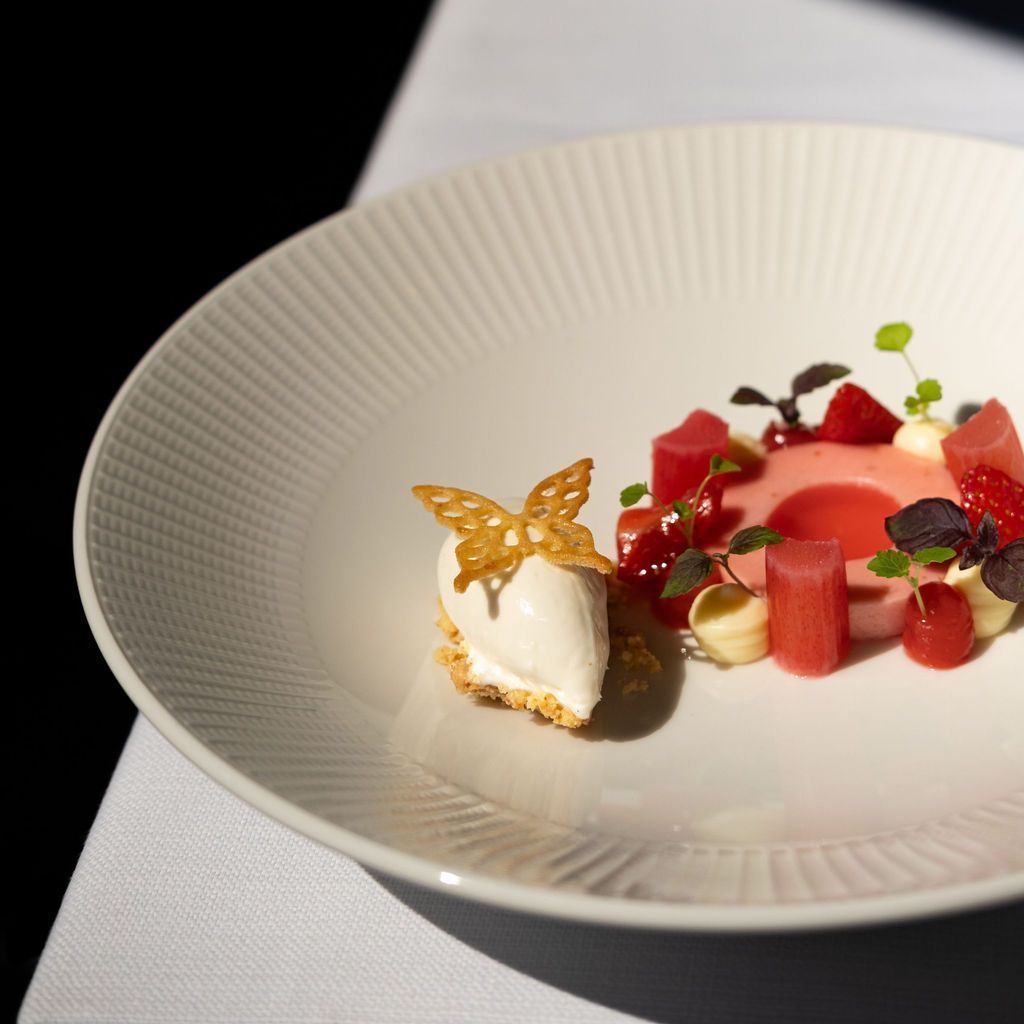 Elegant opgemaakt dessert met witte room, rode fruitblokjes en een gouden vlindergarnering op een wit bord.