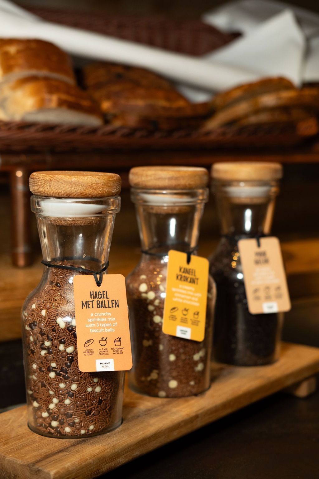 Drie kruidenpotjes met kurken deksels en handgeschreven etiketten op een houten plank in een bakkerijvitrine.