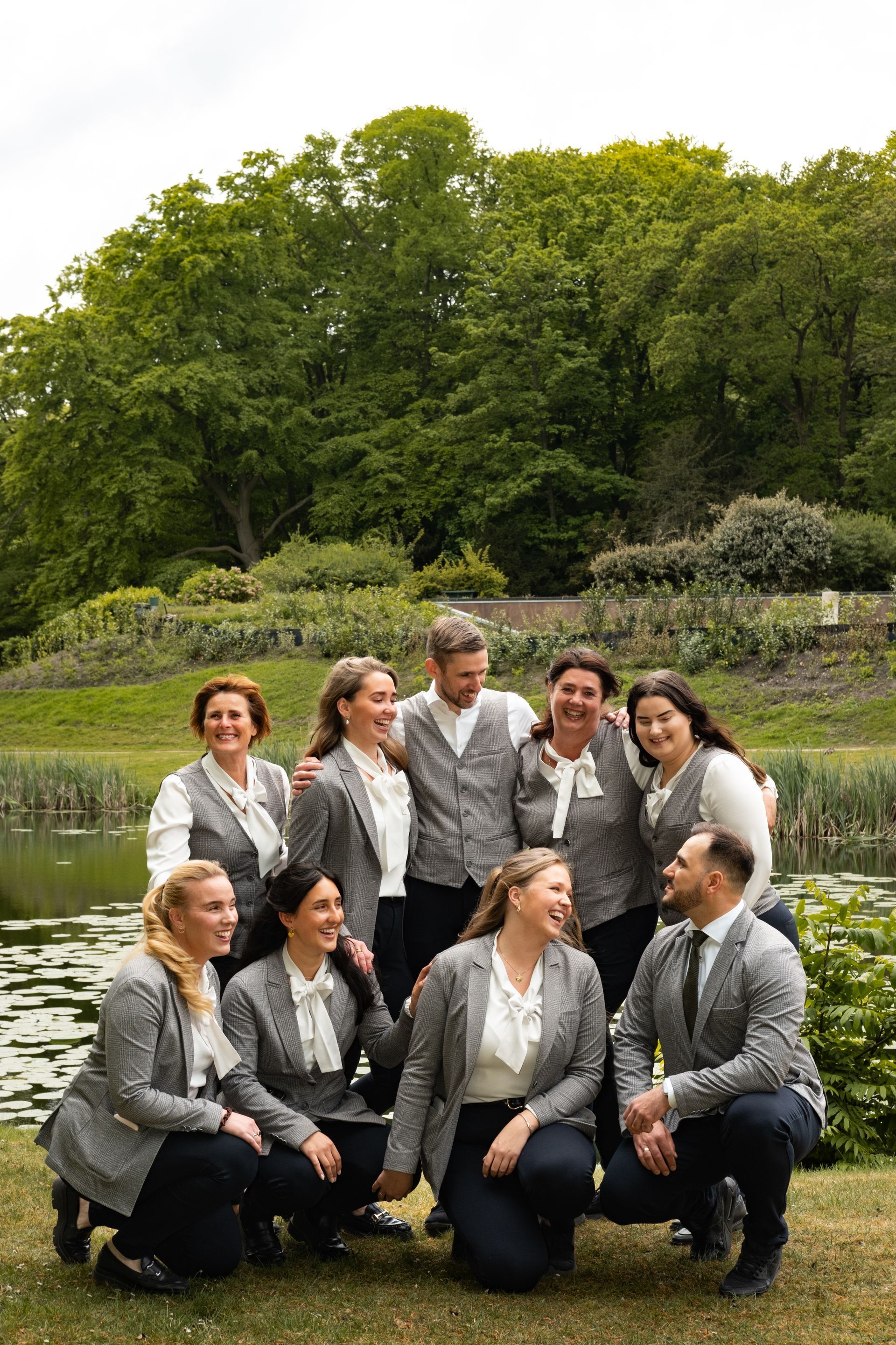 Group of people posing outdoors in front of a pond and green trees, smiling in coordinated gray and black outfits