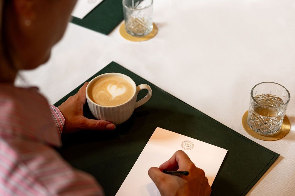 Person writing beside a latte on a green placemat at a white table.
