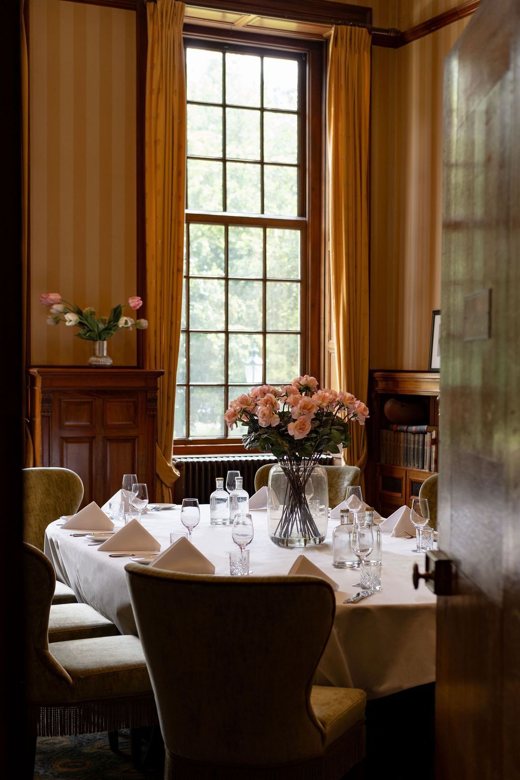 Elegante eetkamer met een gedekte tafel, een bloemstuk in het midden en een hoog raam met warme houten lambrisering.