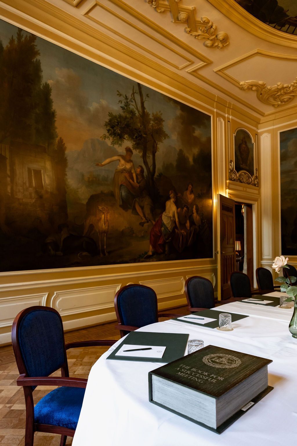 Elegante eetkamer met blauwe stoelen, een wit tafelkleed en een groot ingelijst schilderij aan de muur.