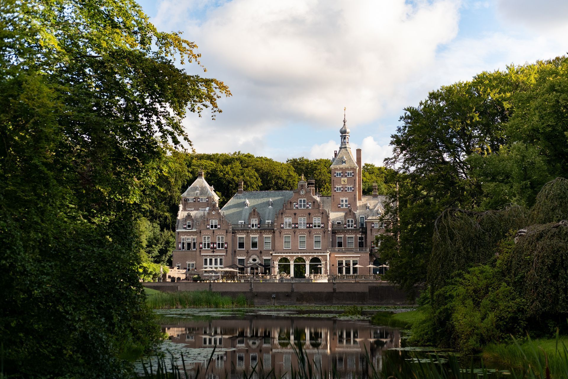 Stone castle-like building with tall tower beside a calm lake, framed by trees and reflected in the water