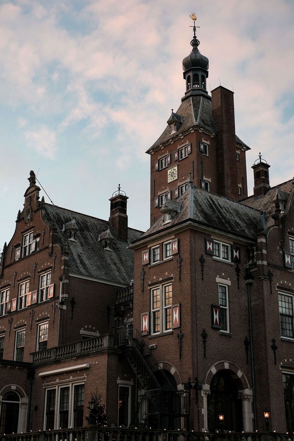 Hohes, historisches Backsteingebäude mit einem zentralen Turm und verzierten Giebeln unter einem bewölkten, rosafarbenen Himmel