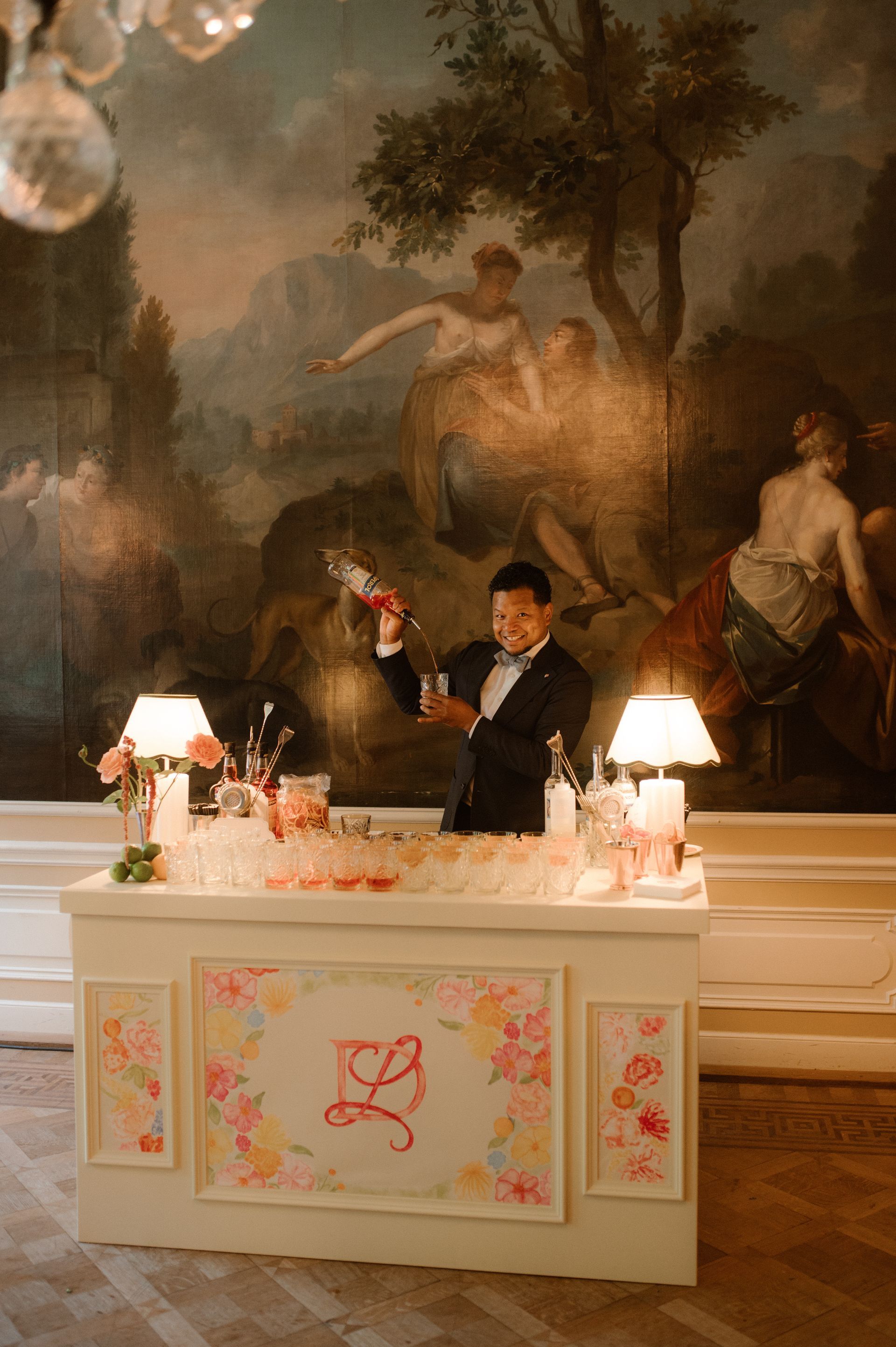 Person seated behind a white floral desk in a formal room with a large mural and table lamps