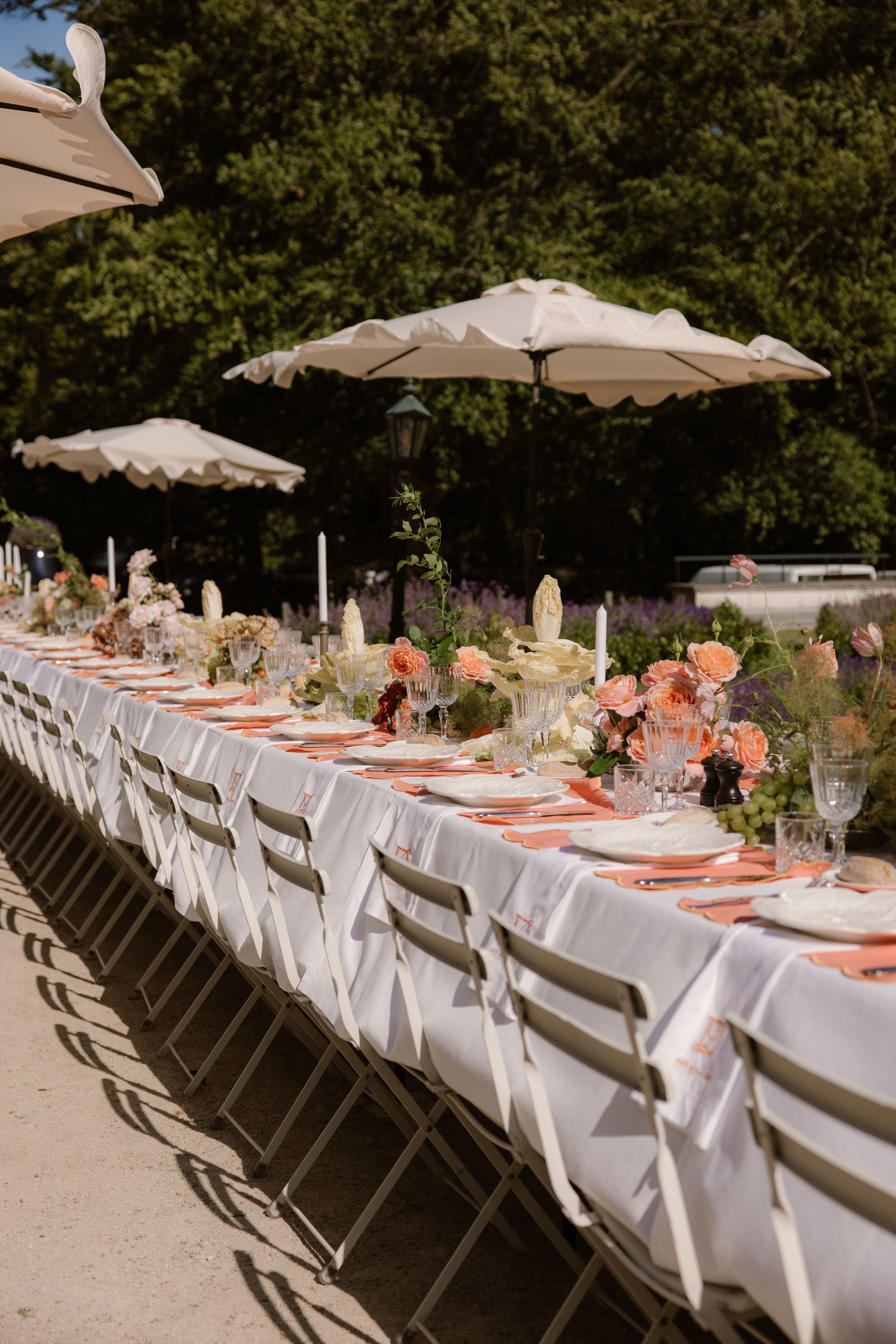Long outdoor banquet table with white chairs, pink floral centerpieces, and umbrellas in a garden setting