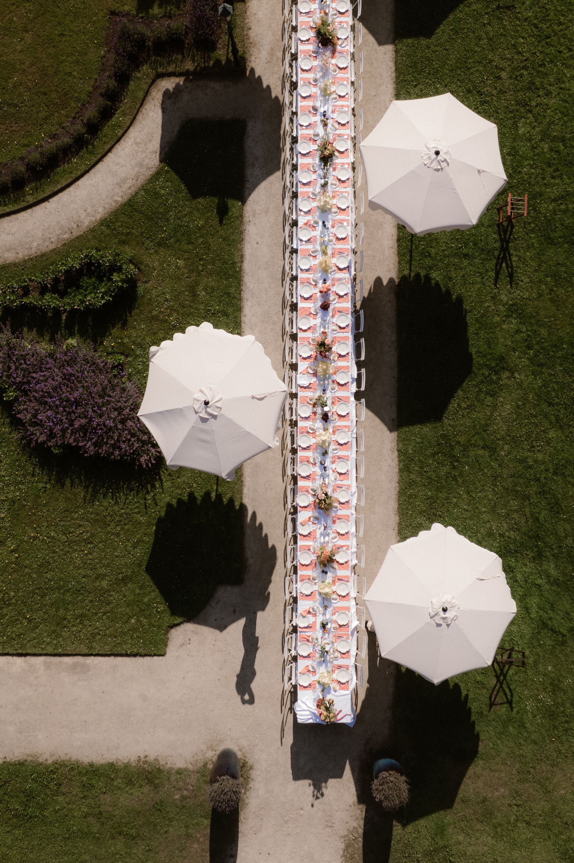 Overhead view of a long banquet table outdoors, lined with white umbrellas and surrounded by greenery.