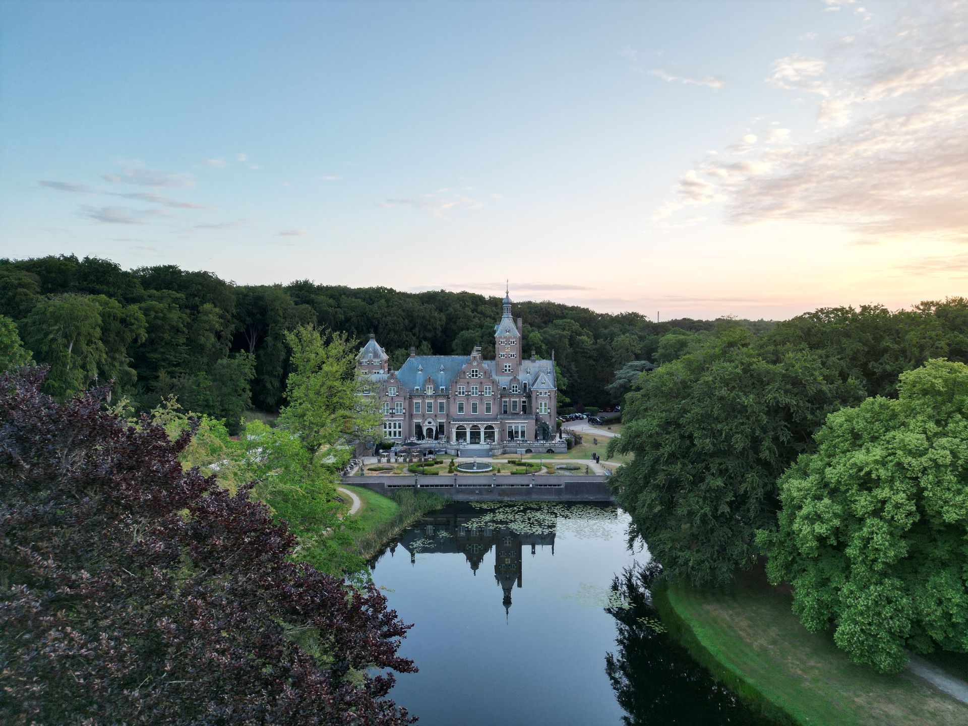 Kasteel aan een spiegelende vijver in de schemering, omgeven door weelderige groene bomen en een pastelkleurige hemel.