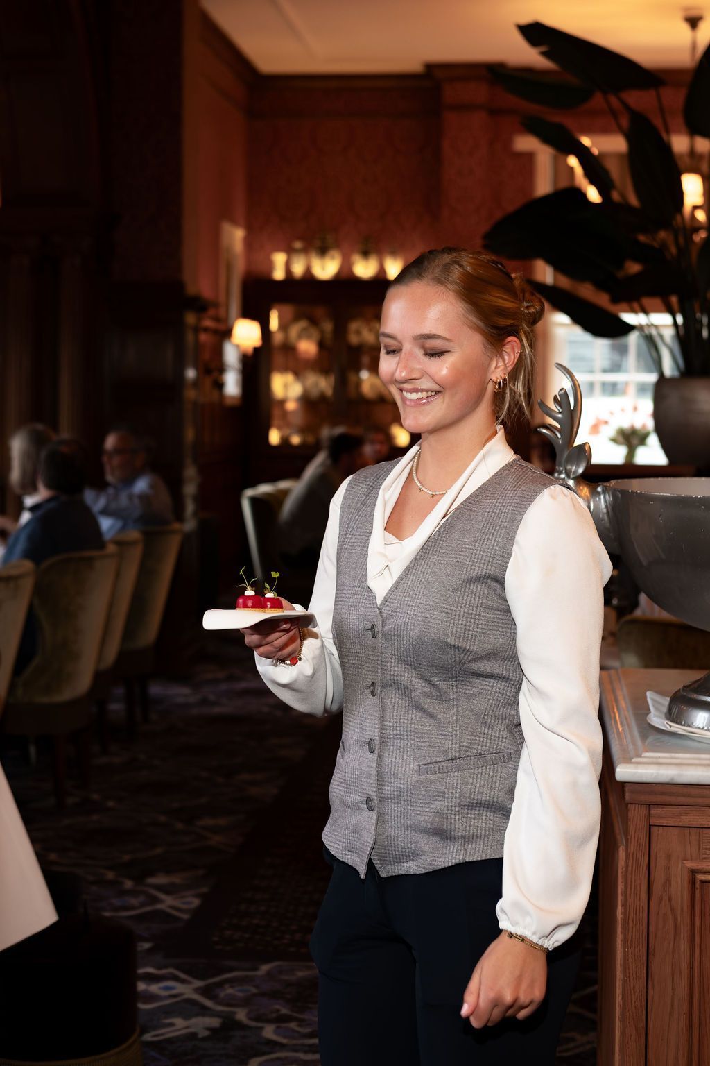 Smiling server in a gray vest holding a dessert in a warmly lit restaurant
