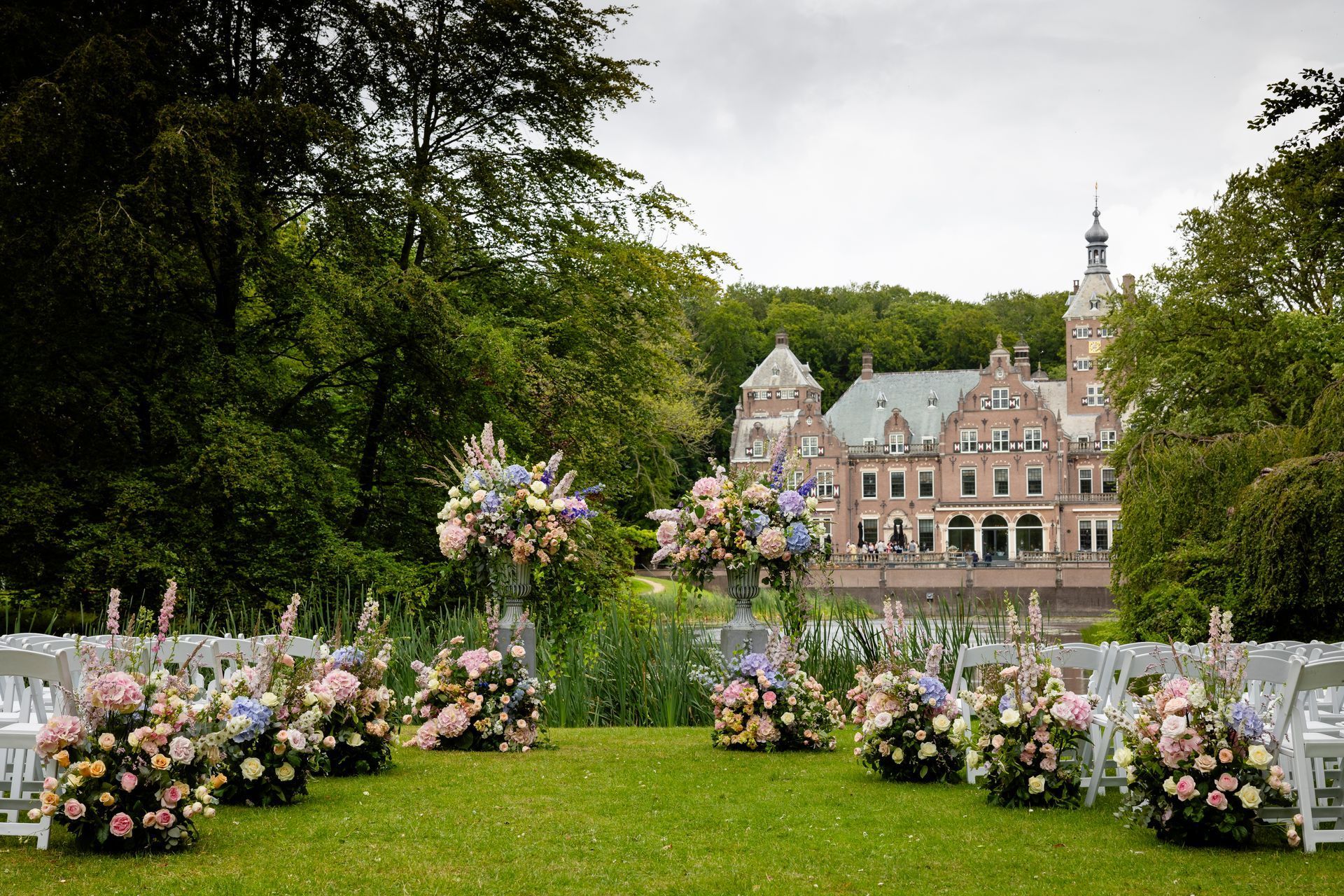 Een bloemenboog op een gazon voor een historisch kasteel, omringd door bomen en pastelkleurige bloemstukken.