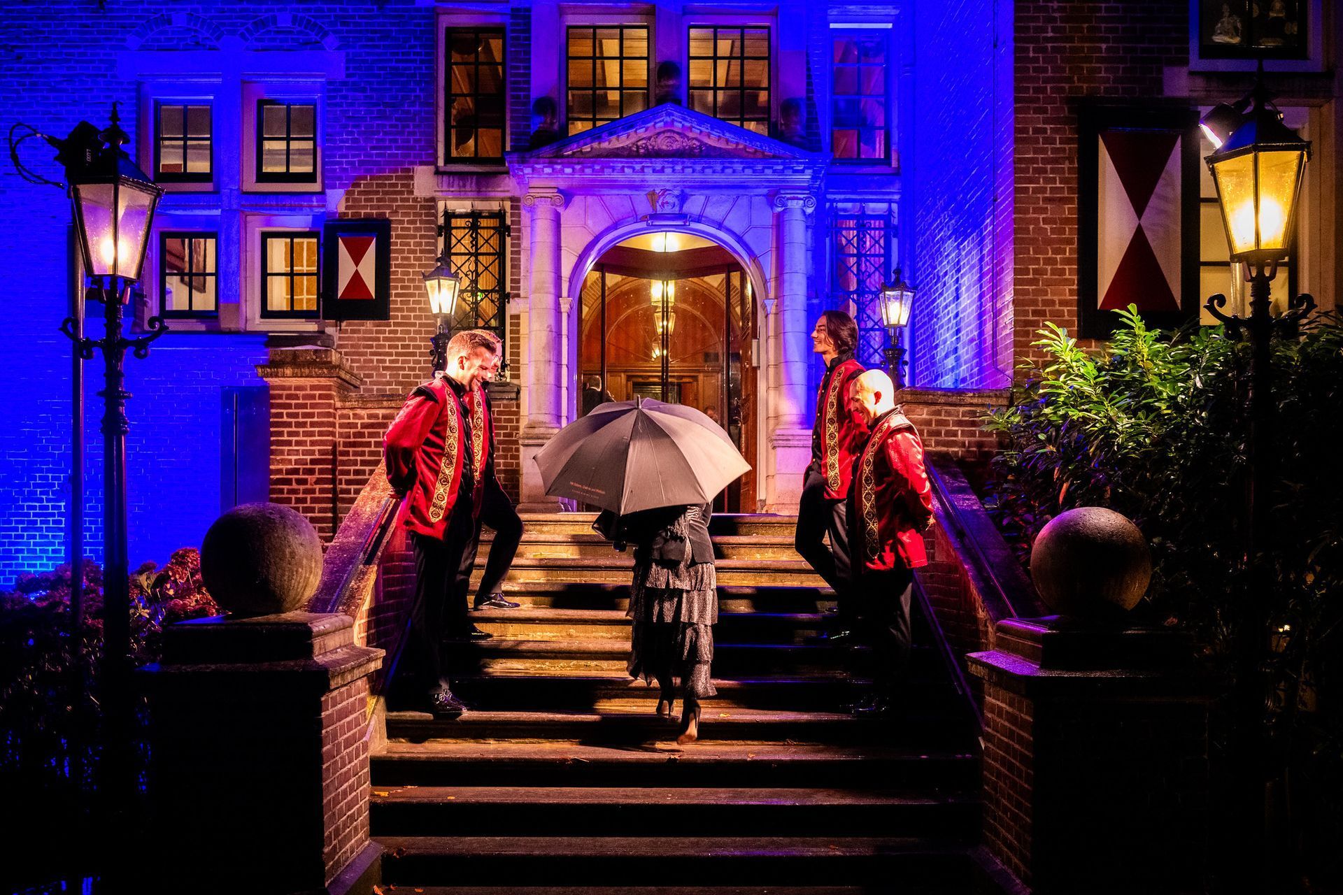 People entering a blue-lit building at night, two holding umbrellas on the steps