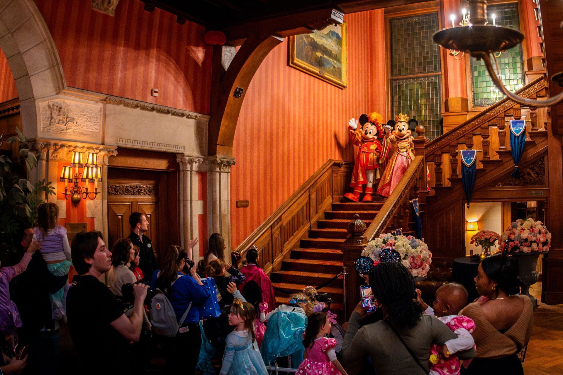 Crowd gathered in a grand wood-paneled hall watching a staircase procession in colorful costumes