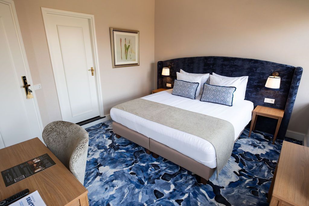 Moderne slaapkamer met donkerblauwe sprei, blauw patroonkleed, bureau en rieten stoel.