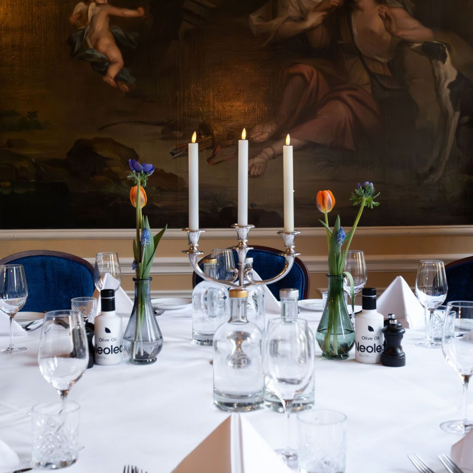 Elegant dining table with white tablecloth, candles, glassware, and small flower arrangements before a painting