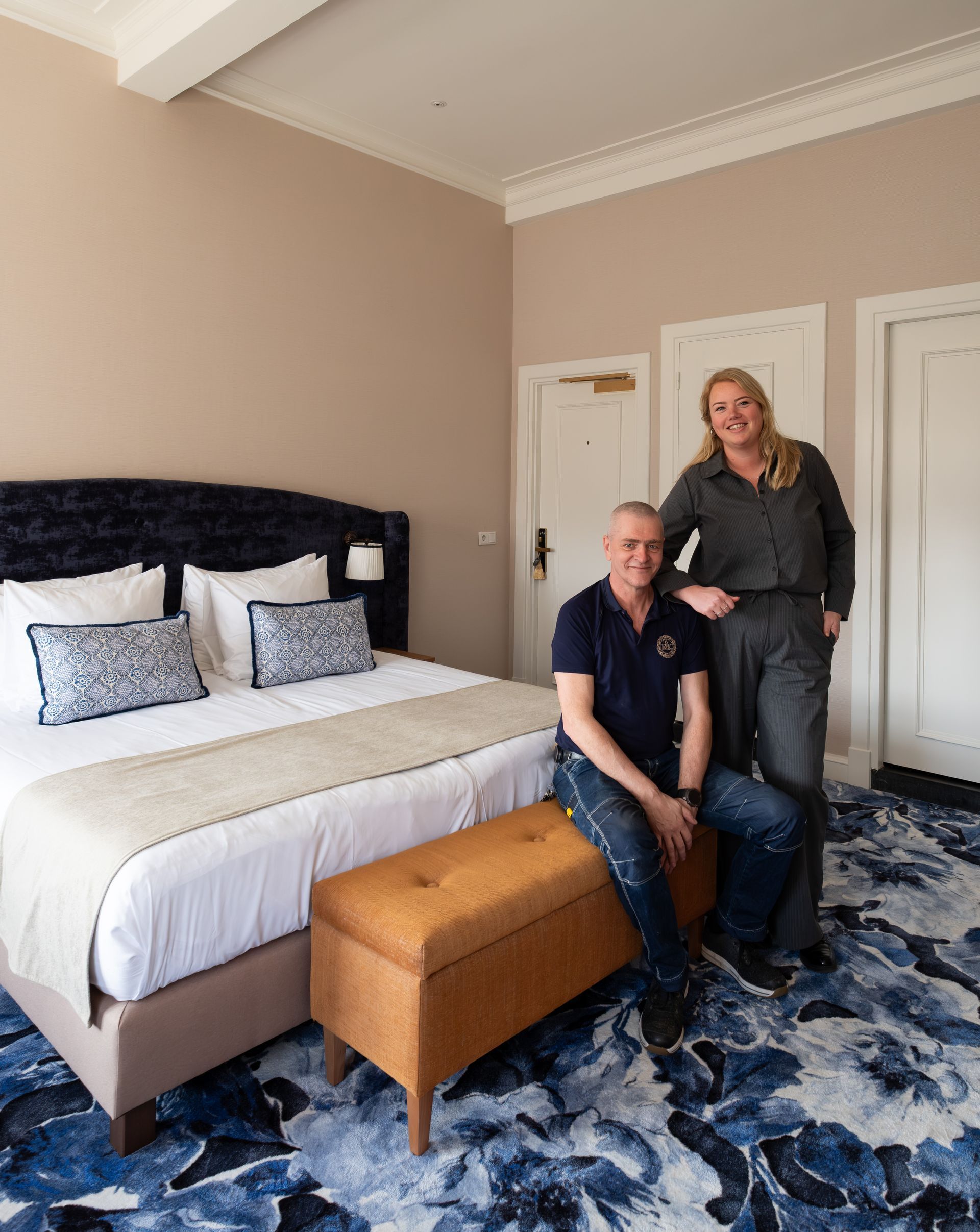 Couple posing in a blue-and-beige bedroom with a bed, bench, and two doors.