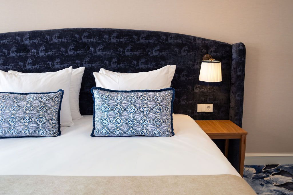 Netjes opgemaakt bed met wit beddengoed en blauwe kussens met patroon, naast een houten nachtkastje en een wandlamp.