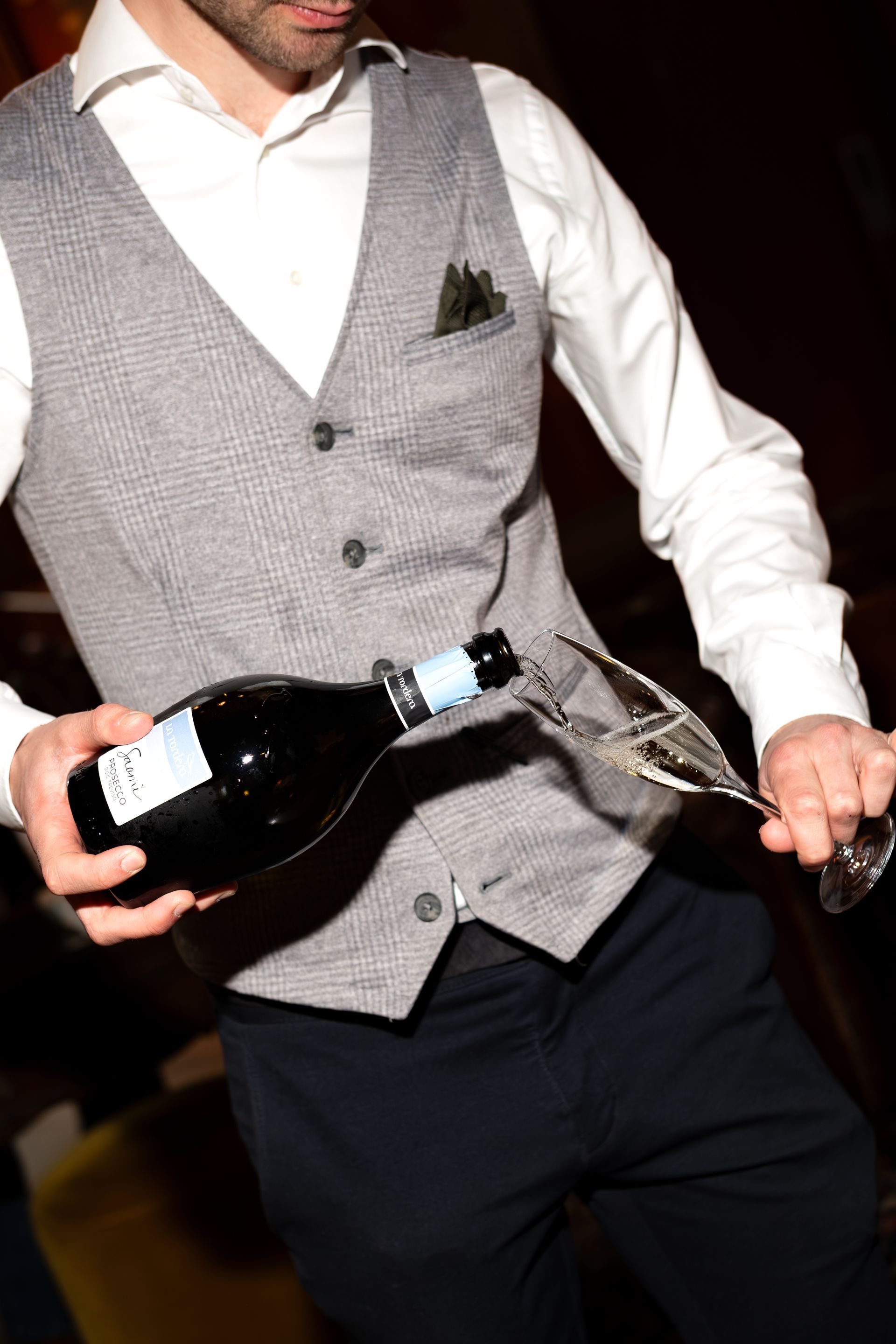 Man in white shirt and gray vest pouring champagne into a glass at a party
