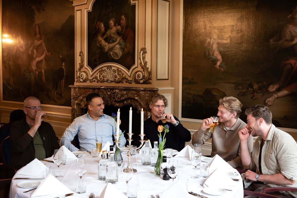 Vijf mensen zitten aan een formele eettafel in een sfeervol verlicht restaurant en praten met elkaar onder het genot van een glas wijn.