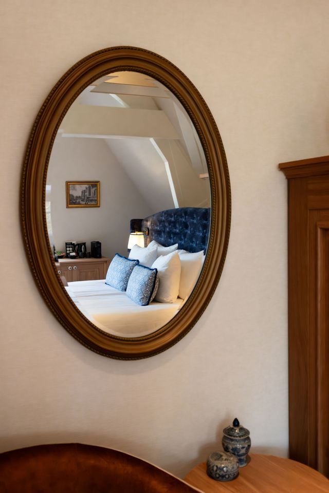 Een ovale spiegel die een gezellige slaapkamer met wit beddengoed en een blauw gecapitonneerd hoofdbord op een crèmekleurige muur weerspiegelt.
