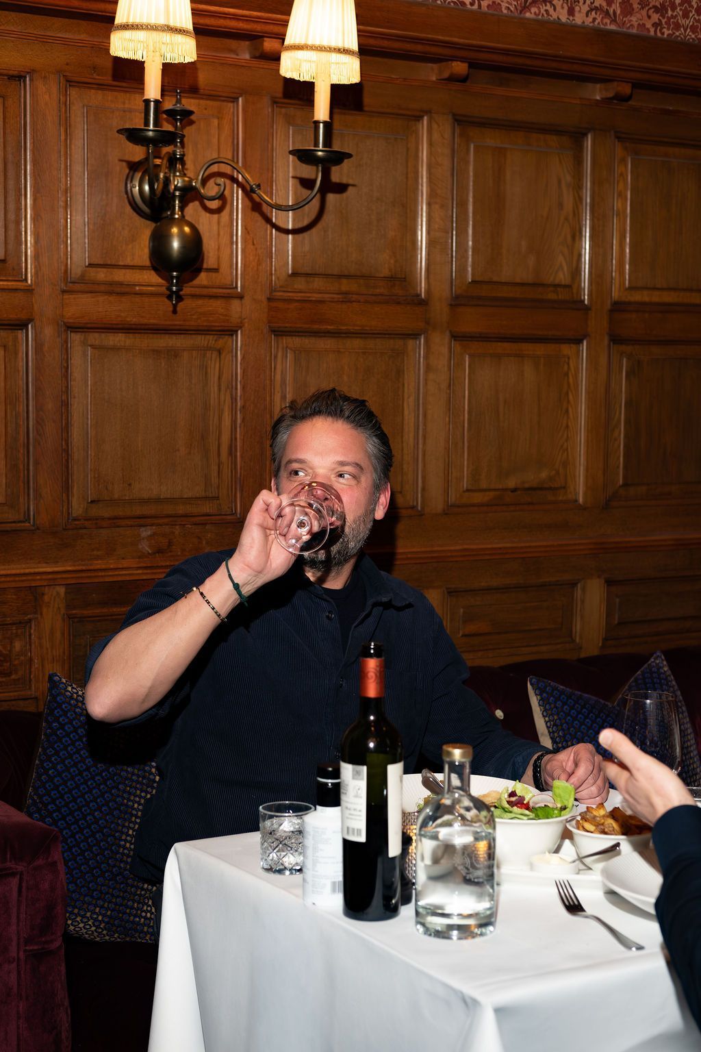 Man drinking from a glass at a restaurant table with wine bottles and a lit wall lamp behind him