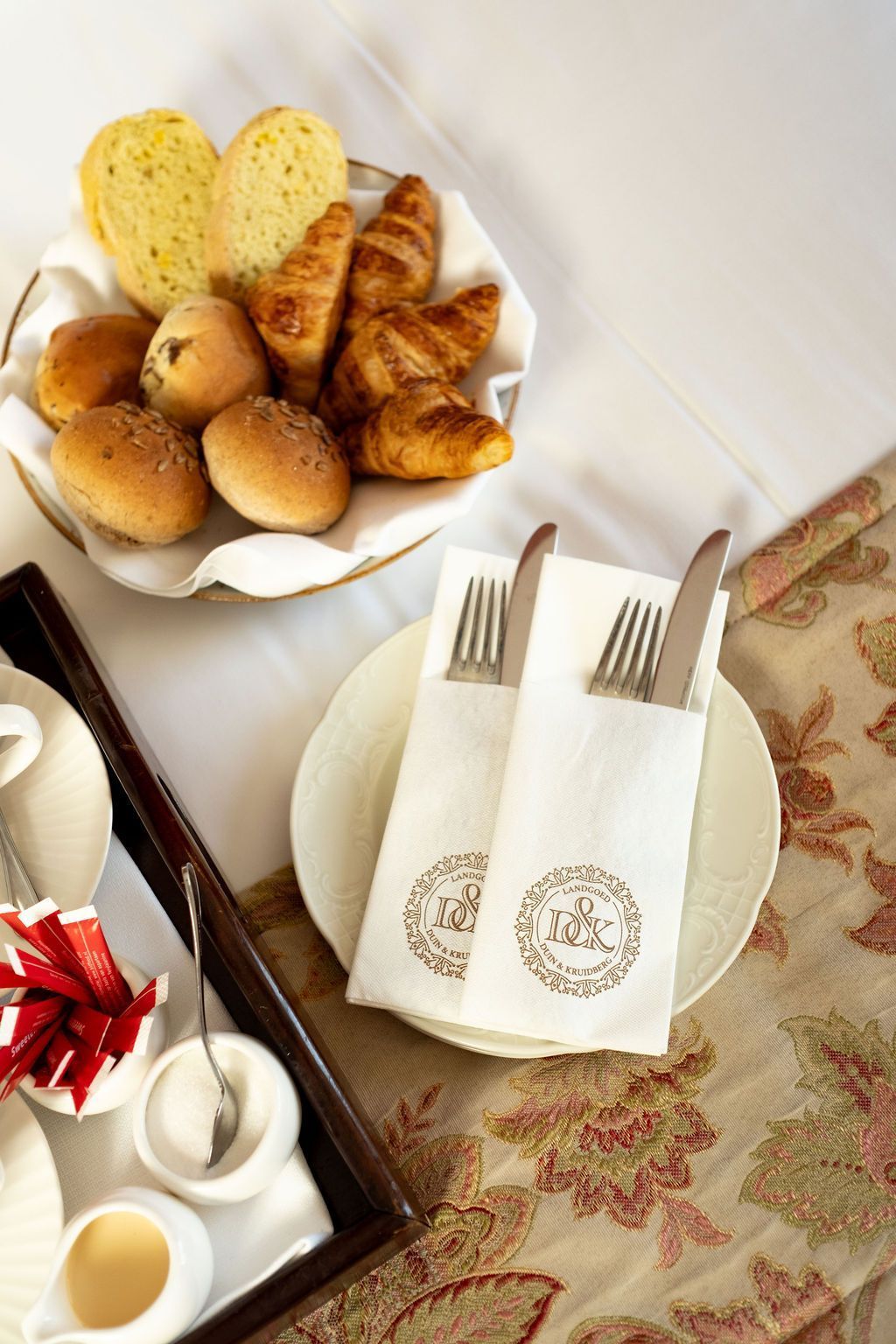 Ontbijttafel met broodjes, croissants, hartvormige toast en gedekte borden met vorken en servetten.