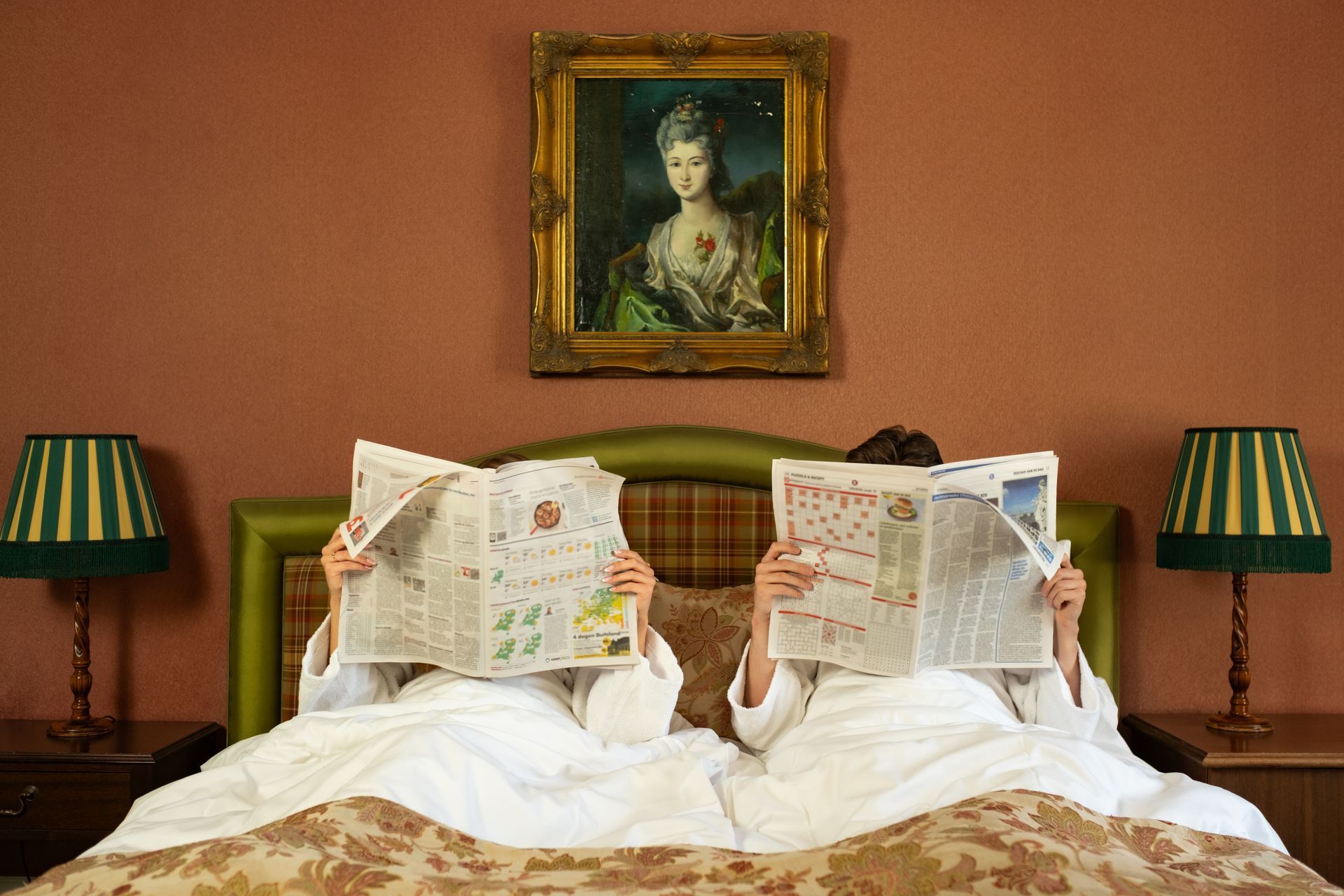 Twee mensen liggen in bed en lezen de krant onder een ingelijst portret, met aan weerszijden bijpassende groene lampen.