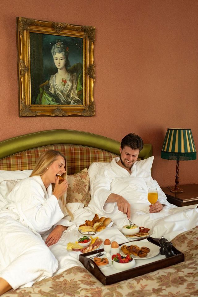 Een stel in witte badjassen ontbijt op een bed in een gezellige hotelkamer.
