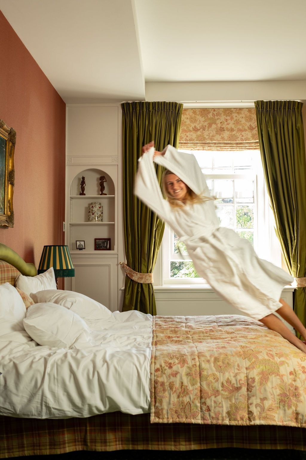Eine Person im weißen Bademantel springt in einem gemütlichen Schlafzimmer mit grünen Vorhängen und Sonnenlicht auf dem Bett herum.