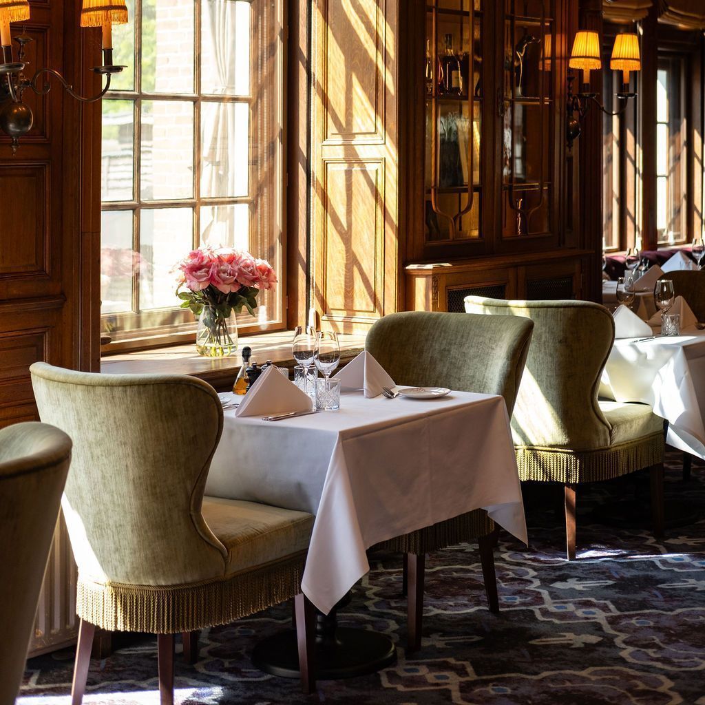 Elegant restaurant dining room with white-tablecloth tables, green chairs, and sunlight streaming through tall windows