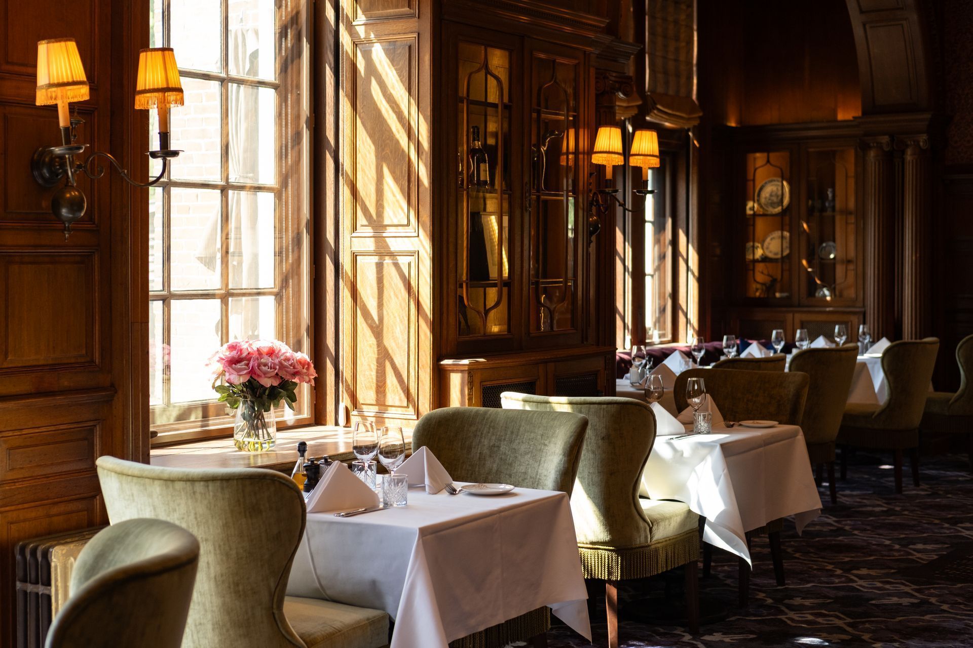 Elegant wood-paneled restaurant with sunlit tables, green chairs, and warm wall lamps