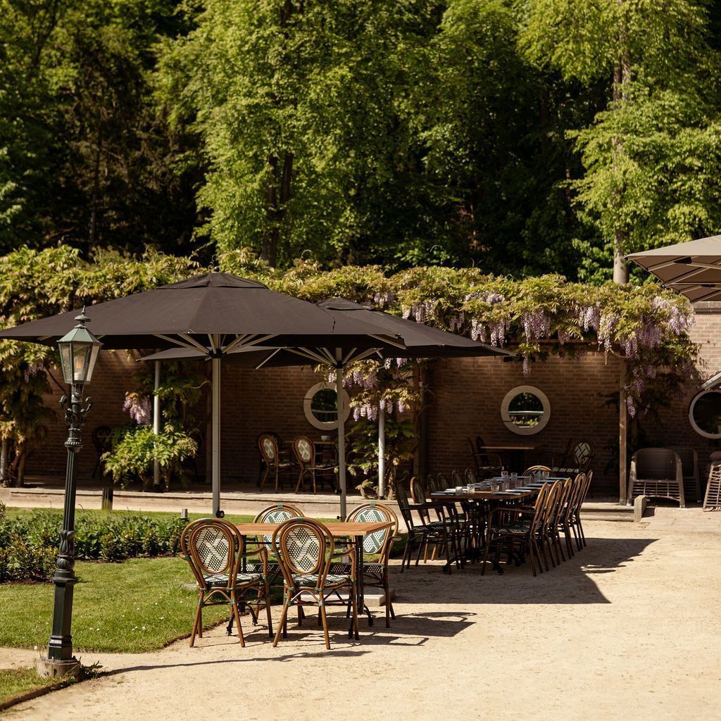 Terras van een café met zwarte parasols, smeedijzeren tafels en stoelen naast een bakstenen gebouw en groen.