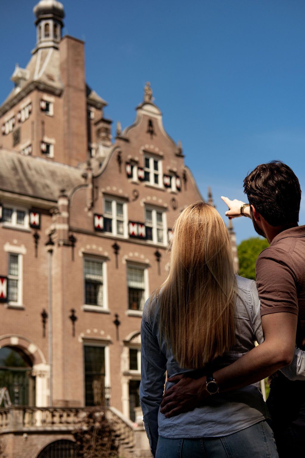 Twee mensen staan ​​arm in arm en wijzen naar een historisch bakstenen gebouw onder een blauwe hemel.